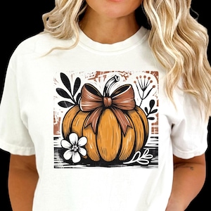 May include: White t-shirt featuring a fall-themed graphic. The design includes an orange pumpkin with a brown bow, surrounded by black leaves and a white flower. The shirt is a comfortable, casual style.