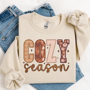 May include: A beige sweatshirt with the text "Cozy season" in brown, orange, and pink letters with a patchwork design. The letters are decorated with fall-themed patterns like leaves, pumpkins, and plaid.