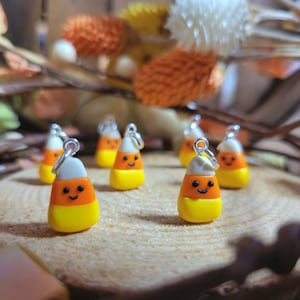 May include: Five small, orange, yellow, and white candy corn charms with smiling faces. The charms are attached to silver jump rings.