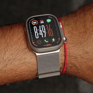 May include: A silver smartwatch with a mesh band displays the time 8:49:16, Tuesday, and other data. The watch has a black screen with white and orange text. A red beaded bracelet is also visible.