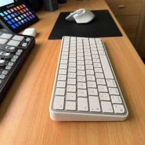 Könnte beinhalten: Eine weiße drahtlose Tastatur mit silbernem Rand. Die Tastatur hat ein Standardlayout mit Funktionstasten, Pfeiltasten und einem Nummernblock. Die Tastatur befindet sich auf einer Holzoberfläche.