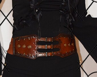 May include: A brown leather belt with a decorative design and three buckles. The belt has a wide, flat design and is adorned with small metal studs.
