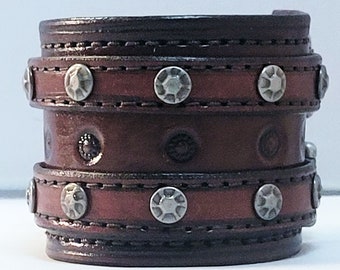 May include: A brown leather cuff bracelet with silver studs. The bracelet has a textured design and is made of multiple layers of leather.