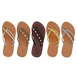 Op de afbeelding: Vijf paar leren teenslippers in verschillende kleuren, elk versierd met decoratieve elementen. De teenslippers hebben verschillende bandjes met schelpen en kralen. De kleuren zijn bruin, zwart, geel en zilver.