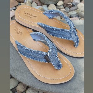 May include: A pair of blue denim flip-flops with brown leather soles. The flip-flops have the text "LESTARIE sandals" on the sole.