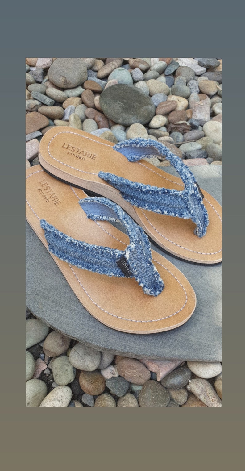 May include: A pair of blue denim flip-flops with brown leather soles. The flip-flops have the text "LESTARIE sandals" on the sole.