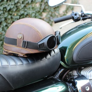 K&ouml;nnte beinhalten: Ein brauner Motorradhelm aus Leder mit gelben N&auml;hten und einer schwarzen Brille liegt auf einem schwarzen Motorradsitz. Der Helm hat einen Vintage-Stil mit einem schwarzen Riemen und einer Schnalle. Das Motorrad ist gr&uuml;n und silber.