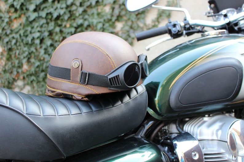 K&ouml;nnte beinhalten: Ein brauner Motorradhelm aus Leder mit gelben N&auml;hten und einer schwarzen Brille liegt auf einem schwarzen Motorradsitz. Der Helm hat einen Vintage-Stil mit einem schwarzen Riemen und einer Schnalle. Das Motorrad ist gr&uuml;n und silber.