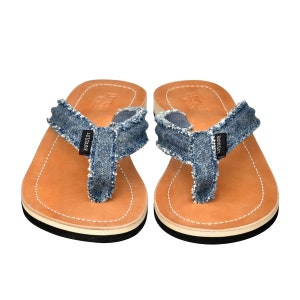 May include: A pair of blue denim flip-flops with brown leather soles and a black trim. The flip-flops have a frayed edge and the brand name "LES TARIE" is printed on the straps.