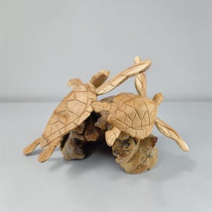May include: A light brown wooden sculpture featuring two sea turtles with carved shell patterns, positioned on a piece of natural driftwood. The turtles have extended flippers and detailed textures, showcasing the natural grain of the wood.