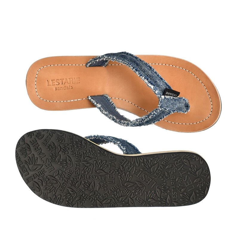 May include: Blue denim flip-flop sandals with a brown leather footbed and a black rubber sole. The sole has the brand name "LESTARIE sandals" printed on it.