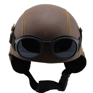 K&ouml;nnte beinhalten: Brauner Helm in Lederoptik mit schwarzer Brille. Der Helm hat gelbe N&auml;hte und Ohrenklappen. Die Brille hat dunkle Gl&auml;ser und einen schwarzen Riemen. Motorrad- oder Fliegerhelm im Vintage-Stil.