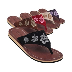 Op de afbeelding: Vijf paar leren sandalen met geborduurde hibiscus bloemen. De sandalen zijn in verschillende kleuren, waaronder bruin, rood, roze, beige en paars.