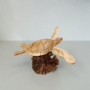 Könnte beinhalten: Eine hellbraune Meeresschildkröten-Skulptur aus Holz mit geschnitztem Panzer-Muster, montiert auf einem dunkelbraunen, strukturierten Stück Treibholz. Die Schildkröte ist mit ausgestreckten Flossen positioniert und zeigt die natürliche Holzmaserung.