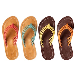 Op de afbeelding: Vier paar leren slippers in verschillende kleuren. De slippers hebben een bruin of bruin voetbed met gekleurde bandjes in rood, teal, geel en oranje. De merknaam "LESTARIE SANDALS" staat op het voetbed.