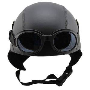 K&ouml;nnte beinhalten: Schwarzer Motorradhelm mit integrierter Brille. Der Helm hat eine runde Form mit einer Naht in der Mitte. Die Brille ist schwarz mit klaren Gl&auml;sern und einem Riemen. Er hat auch Ohrenklappen und einen Kinnriemen.