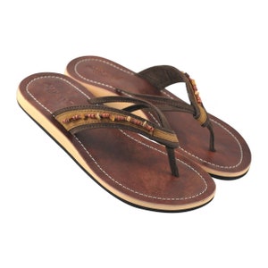 Peut inclure: Sandales en cuir marron à lanières avec une lanière perlée. Les sandales ont une semelle en cuir marron et une lanière en cuir beige.