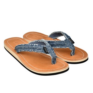 May include: A pair of blue denim flip-flops with brown leather soles and a white trim. The flip-flops have the word "LESSTHANE" printed on the straps.