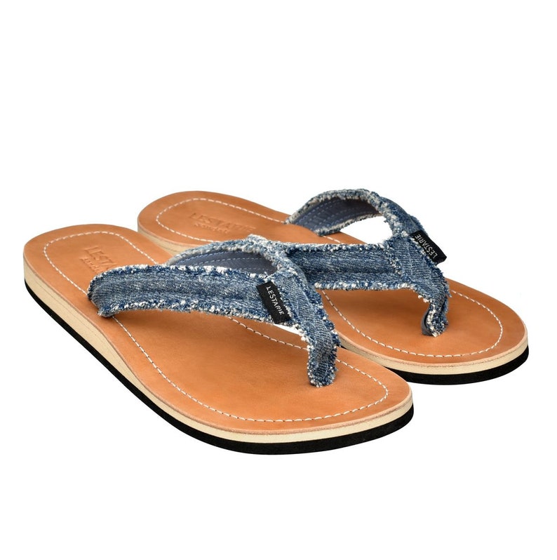 May include: A pair of blue denim flip-flops with brown leather soles and a white trim. The flip-flops have the word "LESSTHANE" printed on the straps.