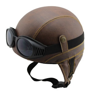 K&ouml;nnte beinhalten: Brauner Motorradhelm aus Leder mit angebrachter schwarzer Brille. Der Helm hat gelbe N&auml;hte und einen schwarzen Riemen zur Befestigung der Brille. Der Helm hat einen Vintage-Stil.