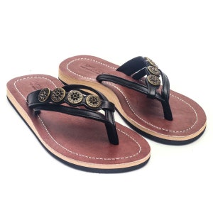 Könnte beinhalten: Ein Paar schwarze Ledersandalen mit brauner Ledersohle. Die Sandalen haben einen Zehensteg und sind mit goldfarbenen Metallelementen verziert.