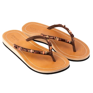 Könnte beinhalten: Braune Leder-Zehentrenner mit einem Perlenband. Die Sandalen haben eine braune Ledersohle und einen kleinen Keilabsatz. Der Markenname "LESDAGUE" ist auf der Sohle aufgedruckt.
