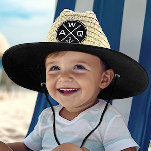 May include: A child's straw hat with a black brim and a circular patch bearing the letters "A", "W", and "Q" with an anchor. The hat has a chin strap. The child is sitting on a blue and white striped beach chair on a sandy beach.