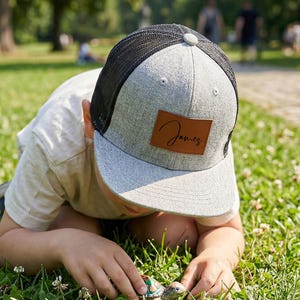 Op de afbeelding: Een grijze en zwarte truckerpet met een bruin leren patch met de naam "James" in cursief. De pet wordt gedragen door een persoon in een park, met groen gras en bomen op de achtergrond.