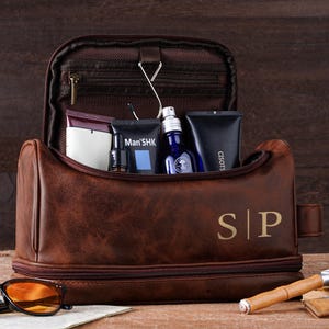 May include: A brown leather wash bag with gold initials "SP". The bag is open, displaying grooming products. Sunglasses and a wooden cigar holder are in the foreground. The bag is on a wooden surface.