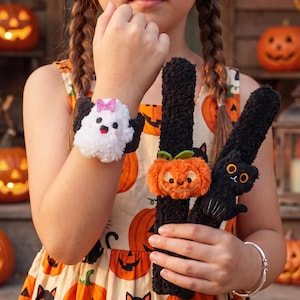 Op de afbeelding: Drie pluche klaparmbanden met Halloween-figuren: een witte geest met een roze glitterstrik, een oranje pompoen met groene blaadjes en een zwarte kat met oranje ogen. Elk figuurtje is bevestigd aan een zwarte, pluizige band.