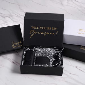 K&ouml;nnte beinhalten: Schwarze Geschenkboxen mit goldfarbenen Schriftz&uuml;gen, darunter eine mit der Aufschrift "Will You Be My Groomsman?" und andere mit Namen. In der ge&ouml;ffneten Box befinden sich eine schwarze Flasche, eine Geldb&ouml;rse und weiteres Zubeh&ouml;r.