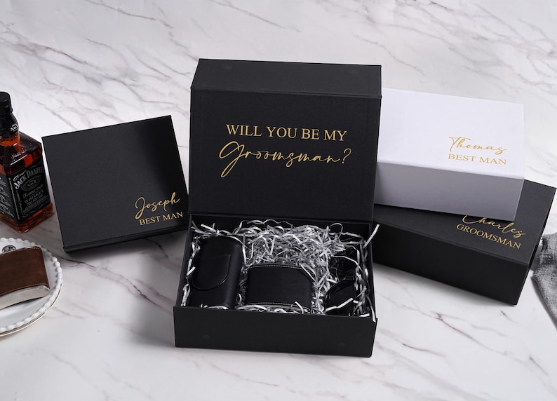 K&ouml;nnte beinhalten: Schwarze Geschenkboxen mit goldfarbenen Schriftz&uuml;gen, darunter eine mit der Aufschrift "Will You Be My Groomsman?" und andere mit Namen. In der ge&ouml;ffneten Box befinden sich eine schwarze Flasche, eine Geldb&ouml;rse und weiteres Zubeh&ouml;r.