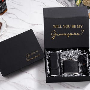 K&ouml;nnte beinhalten: Schwarzes Geschenkbox-Set mit Flachmann, Sonnenbrille und Geldscheinklammer. Die offene Box zeigt den Inhalt und den goldenen Text "WILL YOU BE MY Groomsman?". Eine zweite geschlossene Box tr&auml;gt den Namen "Jackson" und das Wort "Groomsman" in Gold.