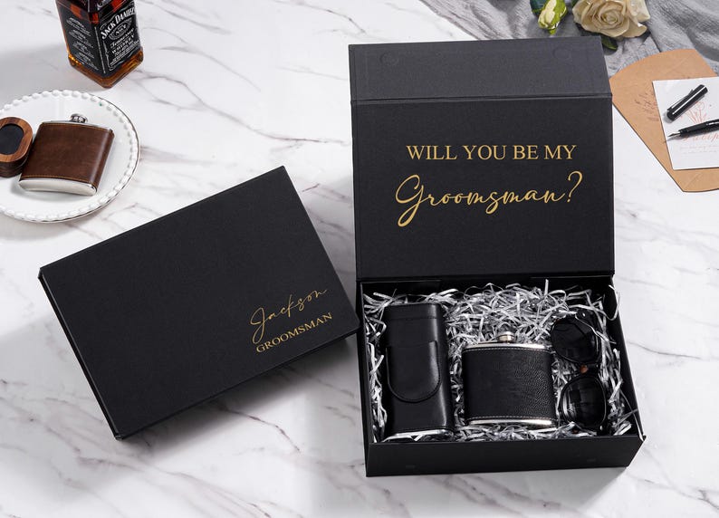 K&ouml;nnte beinhalten: Schwarzes Geschenkbox-Set mit Flachmann, Sonnenbrille und Geldscheinklammer. Die offene Box zeigt den Inhalt und den goldenen Text "WILL YOU BE MY Groomsman?". Eine zweite geschlossene Box tr&auml;gt den Namen "Jackson" und das Wort "Groomsman" in Gold.