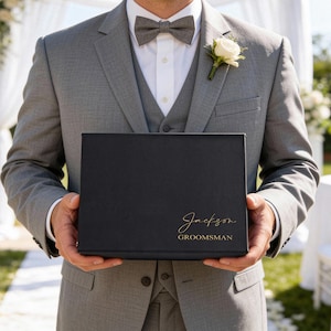 K&ouml;nnte beinhalten: Eine schwarze Geschenkbox mit goldfarbenem Schriftzug "Jackson" und "Groomsman" wird von einer Person in einem grauen Anzug gehalten. Eine wei&szlig;e Rose ist an der Revers des Anzugs befestigt. Der Hintergrund zeigt eine Hochzeitszeremonie.