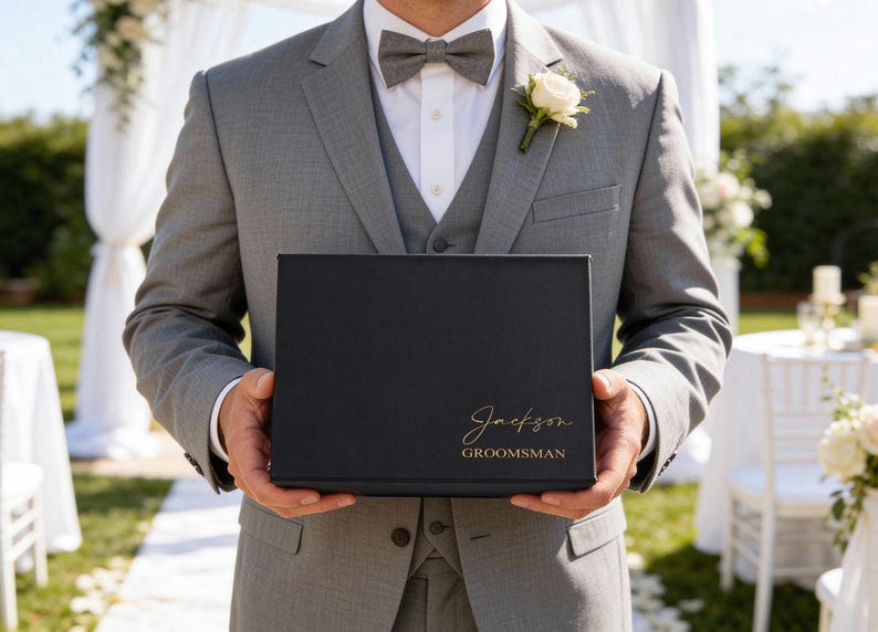 K&ouml;nnte beinhalten: Eine schwarze Geschenkbox mit goldfarbenem Schriftzug "Jackson" und "Groomsman" wird von einer Person in einem grauen Anzug gehalten. Eine wei&szlig;e Rose ist an der Revers des Anzugs befestigt. Der Hintergrund zeigt eine Hochzeitszeremonie.