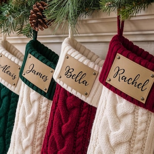 May include: Four cable-knit Christmas stockings in cream, dark green, and deep red. Each stocking features a tan leather-like rectangular patch secured with four metal rivets. The names Alexis, James, Bella, and Rachel are printed in black script.