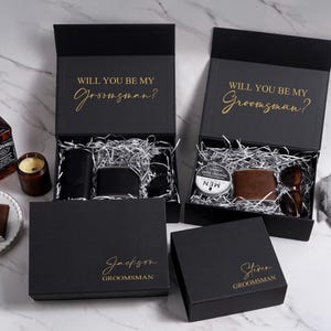 K&ouml;nnte beinhalten: Schwarze Geschenkboxen mit goldenem Text &bdquo;WILL YOU BE MY Groomsman?&ldquo; und personalisierten Namen. Die Boxen enthalten Geldb&ouml;rsen, Sonnenbrillen und Rasiercreme. Eine Whiskyflasche und eine Kerze sind ebenfalls sichtbar.
