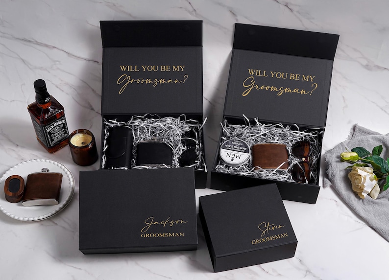 K&ouml;nnte beinhalten: Schwarze Geschenkboxen mit goldenem Text &bdquo;WILL YOU BE MY Groomsman?&ldquo; und personalisierten Namen. Die Boxen enthalten Geldb&ouml;rsen, Sonnenbrillen und Rasiercreme. Eine Whiskyflasche und eine Kerze sind ebenfalls sichtbar.