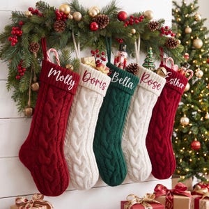 May include: Five cable-knit Christmas stockings in red, white, and green, each featuring a personalized name embroidered in white script: Molly, Doris, Bella, Rose, and Esther. The stockings are hung from a lush evergreen garland with red berries.