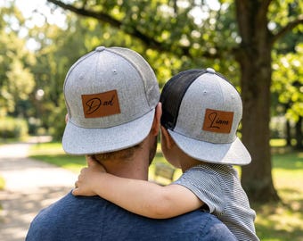 May include: Two gray trucker hats with brown leather patches. One hat has "Dad" and the other has "Liam" printed on the patches. The hats are worn by a person and a child, with the child's arm around the person's neck.