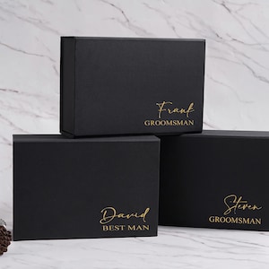 K&ouml;nnte beinhalten: Drei schwarze Geschenkboxen mit goldfarbenen Buchstaben. Auf der oberen Box steht "Frank GROOMSMAN", auf der mittleren Box "David BEST MAN" und auf der unteren Box "Steven GROOMSMAN". Eine Whiskyflasche und ein Teller mit einem Schokoriegel sind im Hintergrund.