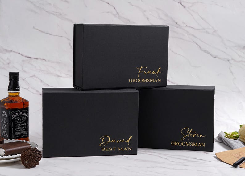 K&ouml;nnte beinhalten: Drei schwarze Geschenkboxen mit goldfarbenen Buchstaben. Auf der oberen Box steht "Frank GROOMSMAN", auf der mittleren Box "David BEST MAN" und auf der unteren Box "Steven GROOMSMAN". Eine Whiskyflasche und ein Teller mit einem Schokoriegel sind im Hintergrund.
