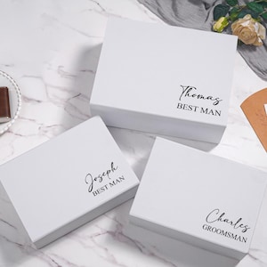 K&ouml;nnte beinhalten: Drei wei&szlig;e Geschenkboxen mit schwarzem Text, der "Thomas BEST MAN", "Joseph BEST MAN" und "Charles GROOMSMAN" lautet. Eine braune Flasche und eine kleine Holzkiste liegen auf einem wei&szlig;en Teller. Die Boxen stehen auf einer wei&szlig;en Marmoroberfl&auml;che.