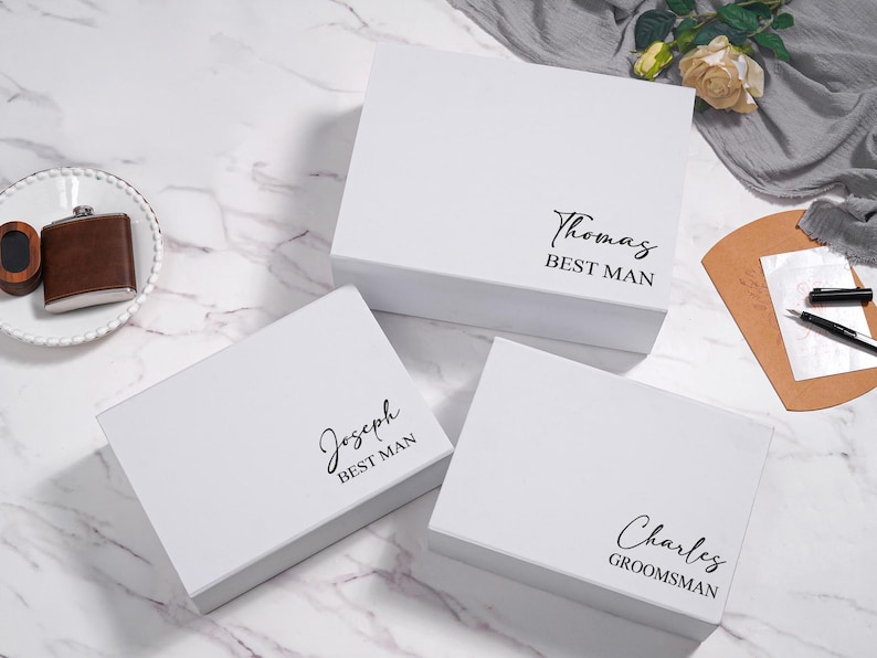 K&ouml;nnte beinhalten: Drei wei&szlig;e Geschenkboxen mit schwarzem Text, der "Thomas BEST MAN", "Joseph BEST MAN" und "Charles GROOMSMAN" lautet. Eine braune Flasche und eine kleine Holzkiste liegen auf einem wei&szlig;en Teller. Die Boxen stehen auf einer wei&szlig;en Marmoroberfl&auml;che.