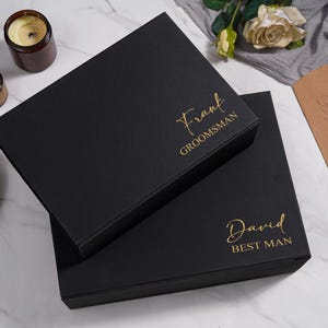 K&ouml;nnte beinhalten: Zwei schwarze Geschenkboxen mit goldfarbener Aufschrift "Frank Groomsman" und "David Best Man". Die Boxen stehen auf einer Marmoroberfl&auml;che, im Hintergrund Kerzen, eine Flasche und eine Spirituosenflasche.