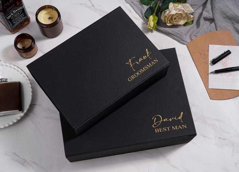 K&ouml;nnte beinhalten: Zwei schwarze Geschenkboxen mit goldfarbener Aufschrift "Frank Groomsman" und "David Best Man". Die Boxen stehen auf einer Marmoroberfl&auml;che, im Hintergrund Kerzen, eine Flasche und eine Spirituosenflasche.