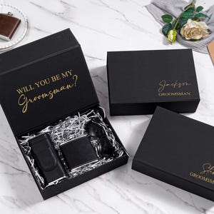 K&ouml;nnte beinhalten: Schwarze Geschenkboxen mit goldfarbenem Schriftzug. Eine Box ist ge&ouml;ffnet und zeigt eine Flasche, eine Sonnenbrille und ein Feuerzeug. Auf dem offenen Deckel steht "WILL YOU BE MY Groomsman?". Zwei weitere Boxen sind geschlossen.