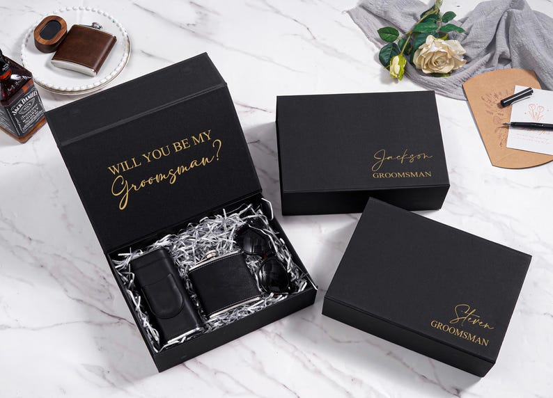 K&ouml;nnte beinhalten: Schwarze Geschenkboxen mit goldfarbenem Schriftzug. Eine Box ist ge&ouml;ffnet und zeigt eine Flasche, eine Sonnenbrille und ein Feuerzeug. Auf dem offenen Deckel steht "WILL YOU BE MY Groomsman?". Zwei weitere Boxen sind geschlossen.