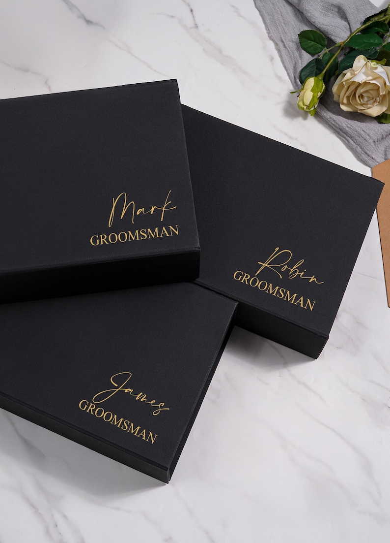 K&ouml;nnte beinhalten: Drei schwarze Geschenkboxen mit goldfarbenen Schriftz&uuml;gen. Die Boxen tragen die Aufschriften "Mark GROOMSMAN", "Robin GROOMSMAN" und "James GROOMSMAN". Die Boxen sind auf einer wei&szlig;en Marmoroberfl&auml;che angeordnet.